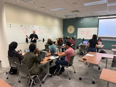 A member works through the Bell state math on a whiteboard covered in red equations while the room follows along at their laptops.