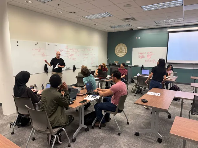 A member works through the Bell state math on a whiteboard covered in red equations while the room follows along at their laptops.
