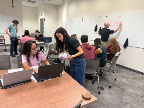 A member holds two glowing Qubis while helping another member at a laptop; behind them someone writes probability math on the whiteboard.
