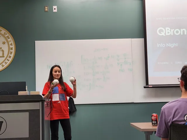 A presenter holds up two white handheld devices in front of a whiteboard full of qubit states, next to the QBronco info night slide.