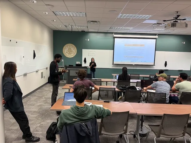 A presenter at the front of the room talks through the project plan on the projector while students listen from their laptops.