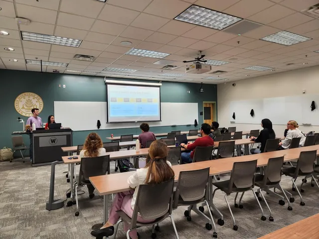 A full classroom facing two presenters at the podium, with a slide that reads: Meeting weekly: Study, Lab, Repeat.