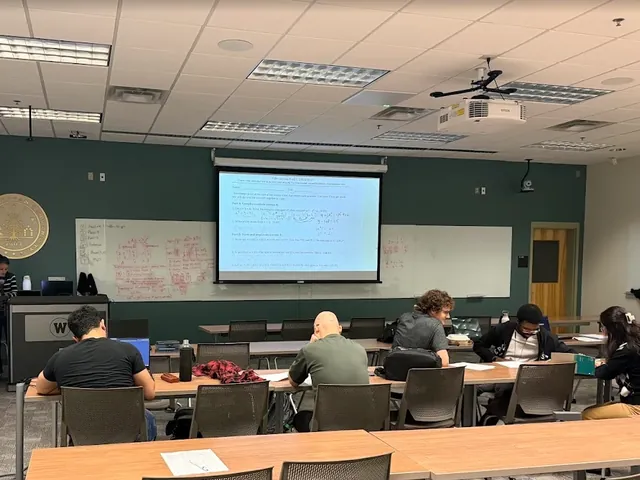 Students working through a worksheet at long desks, with whiteboards covered in red math behind them.